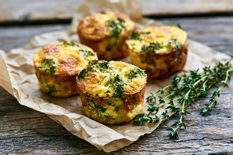 Four broccoli cheddar egg muffins arranged on crinkled parchment paper on a rustic wooden table with fresh thyme sprig
