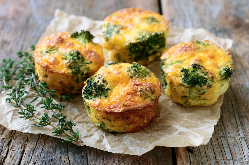 Four broccoli cheddar egg muffins arranged on crinkled parchment paper on a wooden surface with fresh thyme garnish