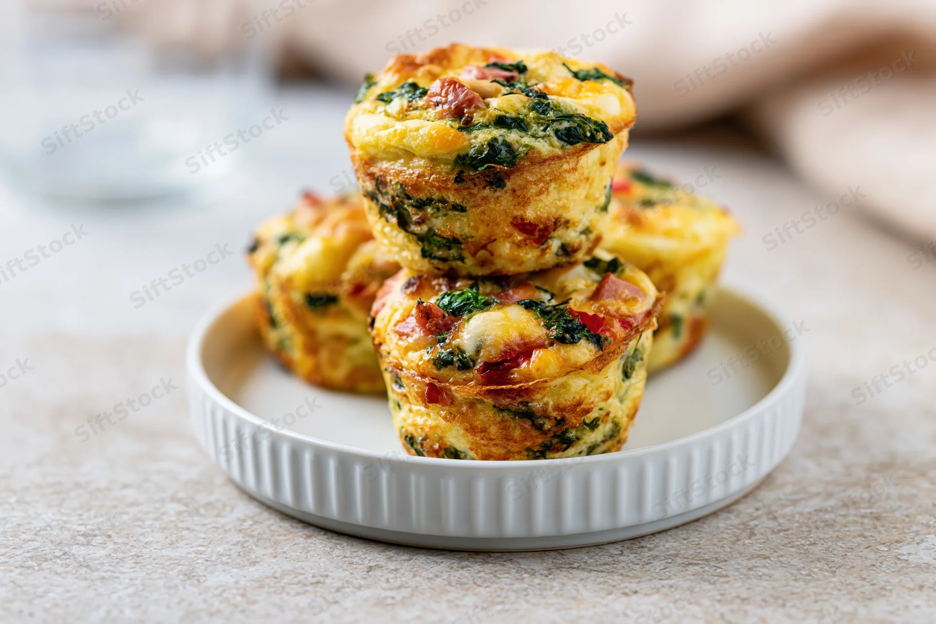 Golden-brown egg muffins with spinach and bacon stacked on a small white ridged plate on a textured countertop