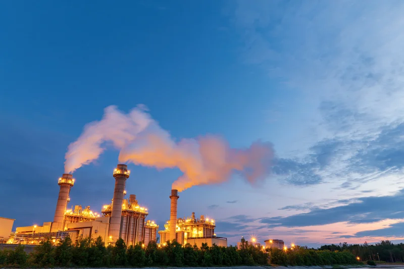Gas turbine power plant emitting steam at dusk with illuminated structures and surrounding greenery