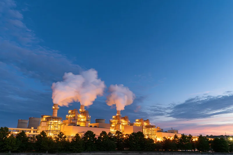 Gas turbine power plant glowing with warm amber lights at dusk against a blue sky with steam rising from smokestacks