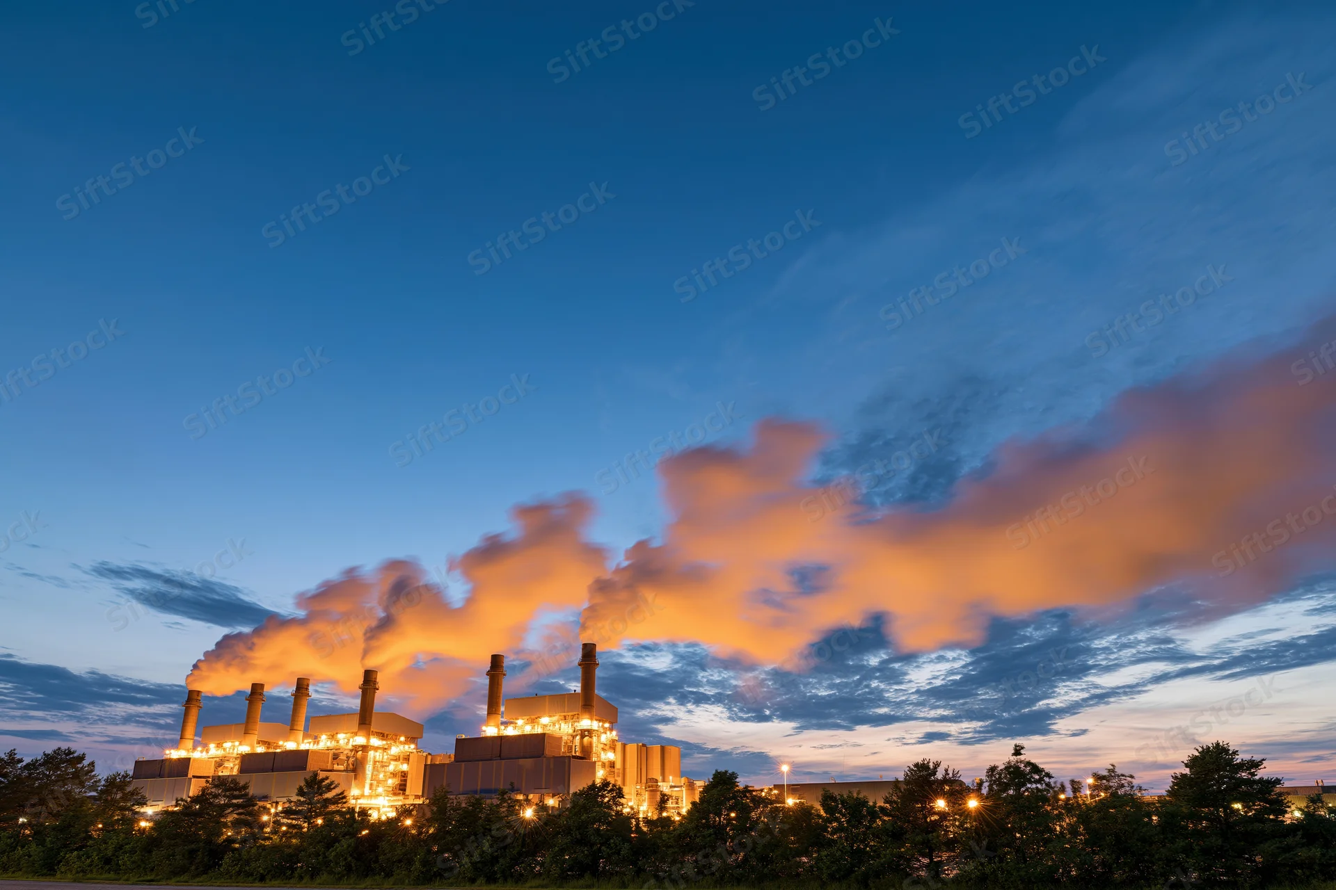Gas turbine power plant glowing with warm amber lights at dusk against a twilight sky with orange clouds and smoke emissions