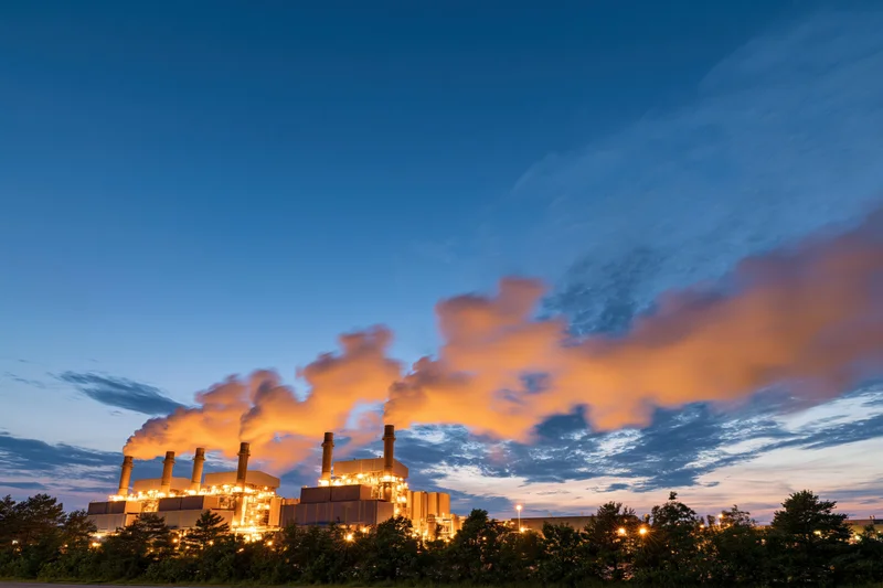 Gas turbine power plant glowing with warm amber lights at dusk against a twilight sky with orange clouds and smoke emissions