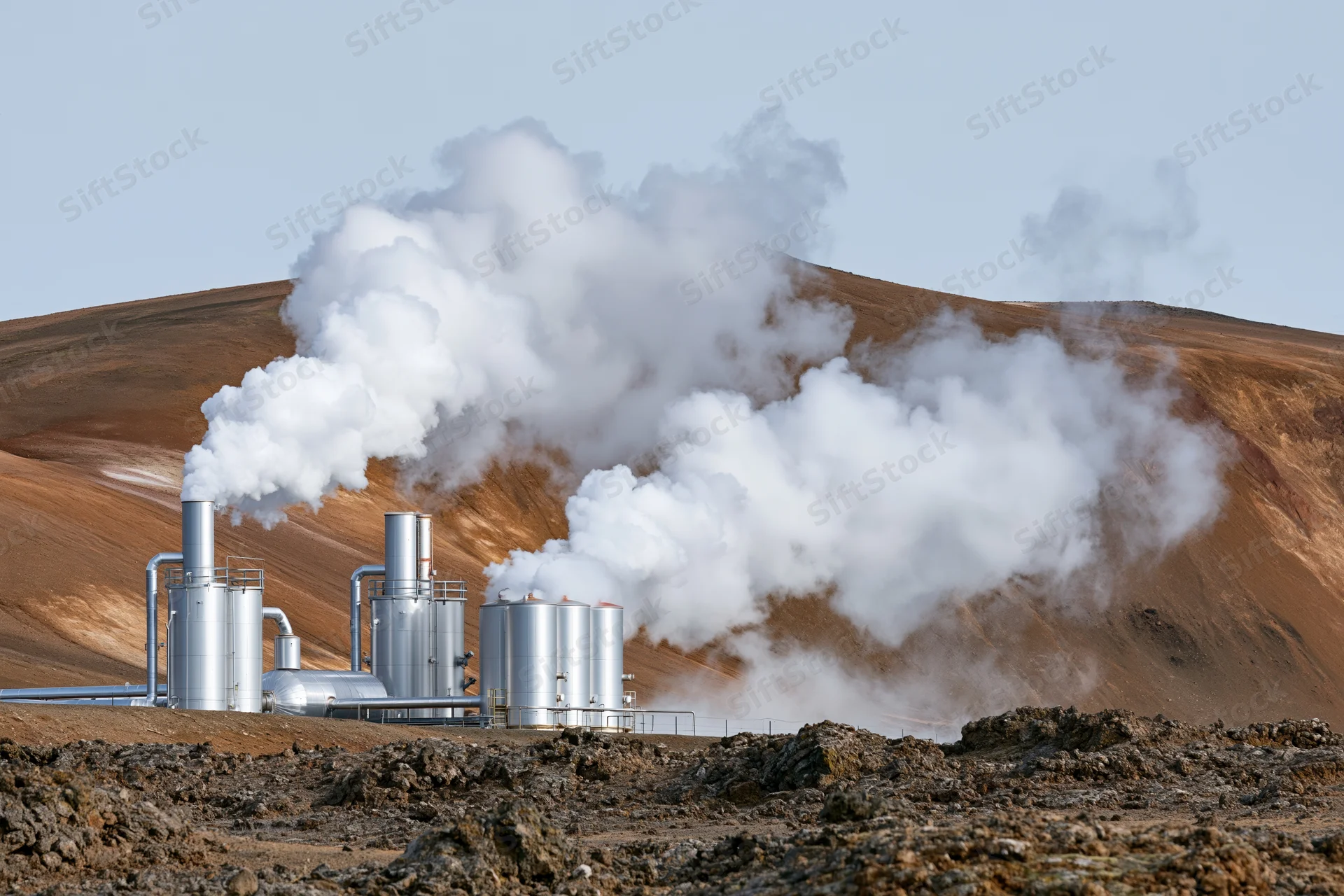 Geothermal power plant with silver metal tanks and stacks releasing steam in arid rocky landscape