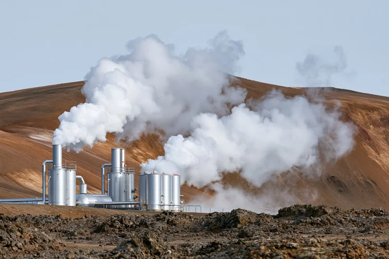 Geothermal power plant with silver metal tanks and stacks releasing steam in arid rocky landscape