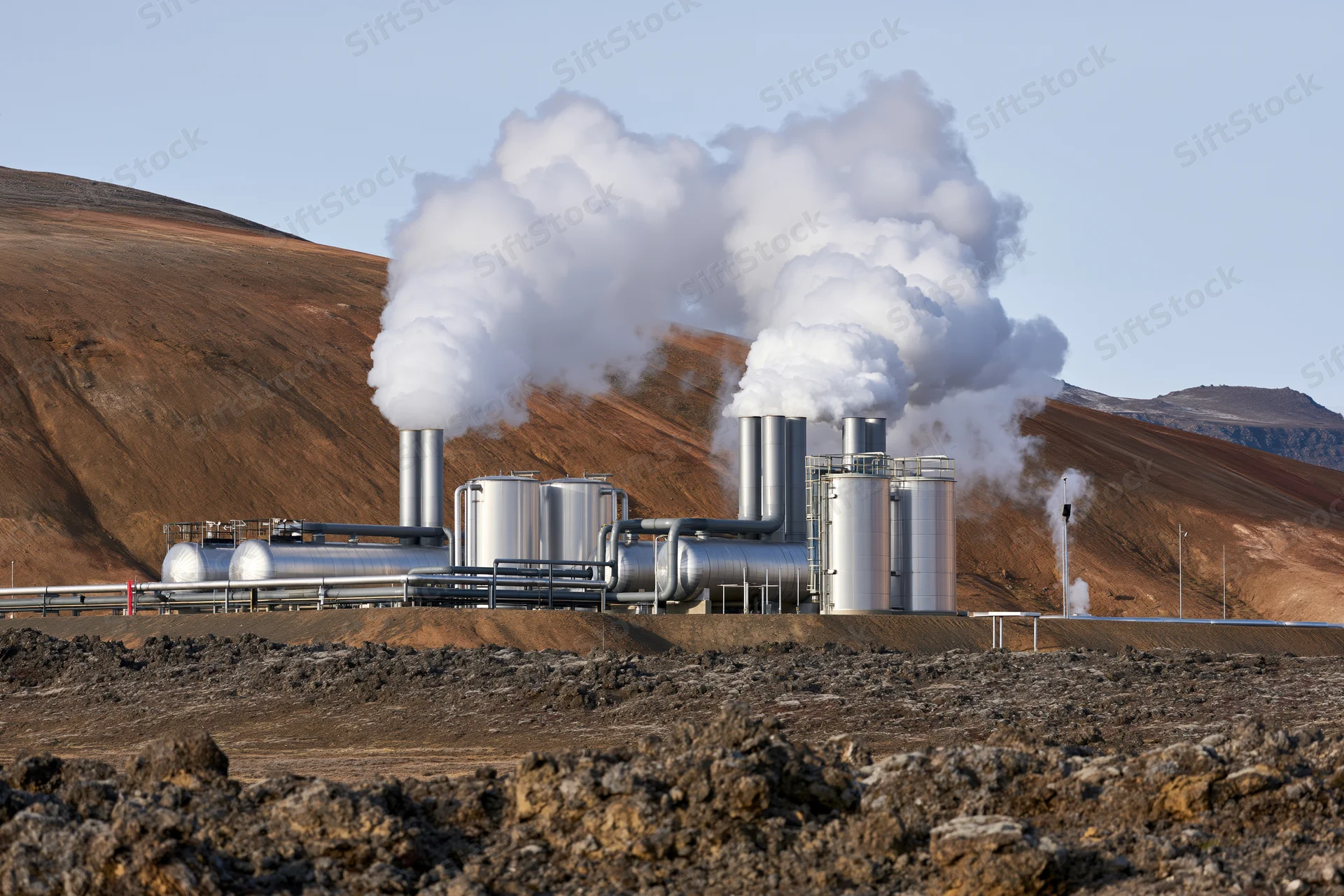 Geothermal power plant with silver metal tanks and stacks emitting steam in arid rocky landscape