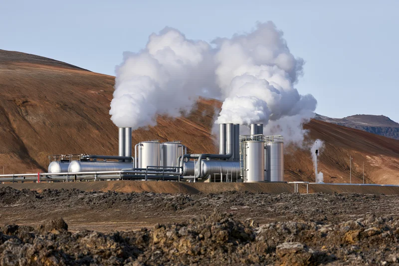 Geothermal power plant with silver metal tanks and stacks emitting steam in arid rocky landscape
