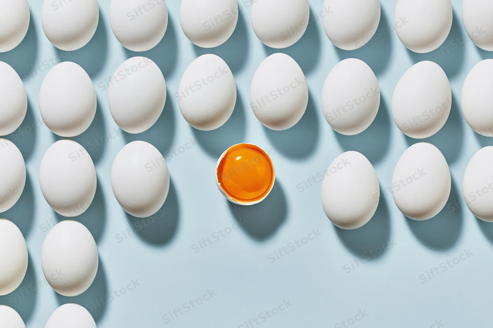 Grid of white eggs arranged in uniform rows on pale blue background with one cracked egg showing orange yolk in center
