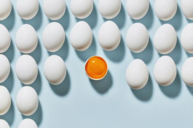 Grid of white eggs arranged in uniform rows on pale blue background with one cracked egg showing orange yolk in center