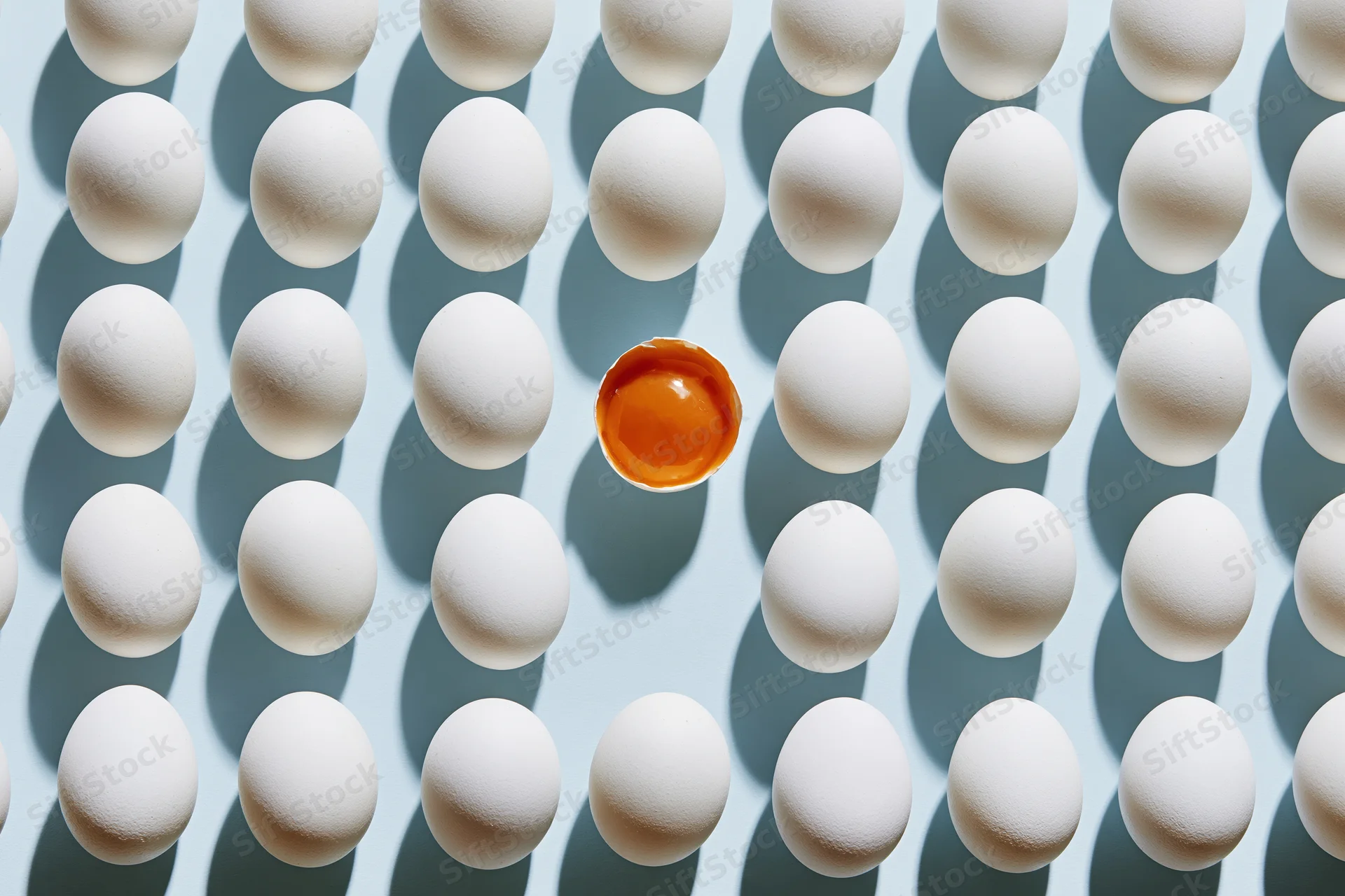 Grid of white eggs arranged in uniform rows with one cracked egg revealing orange yolk at center on pale blue background