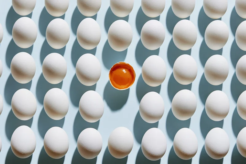 Grid of white eggs arranged in uniform rows with one cracked egg revealing orange yolk at center on pale blue background