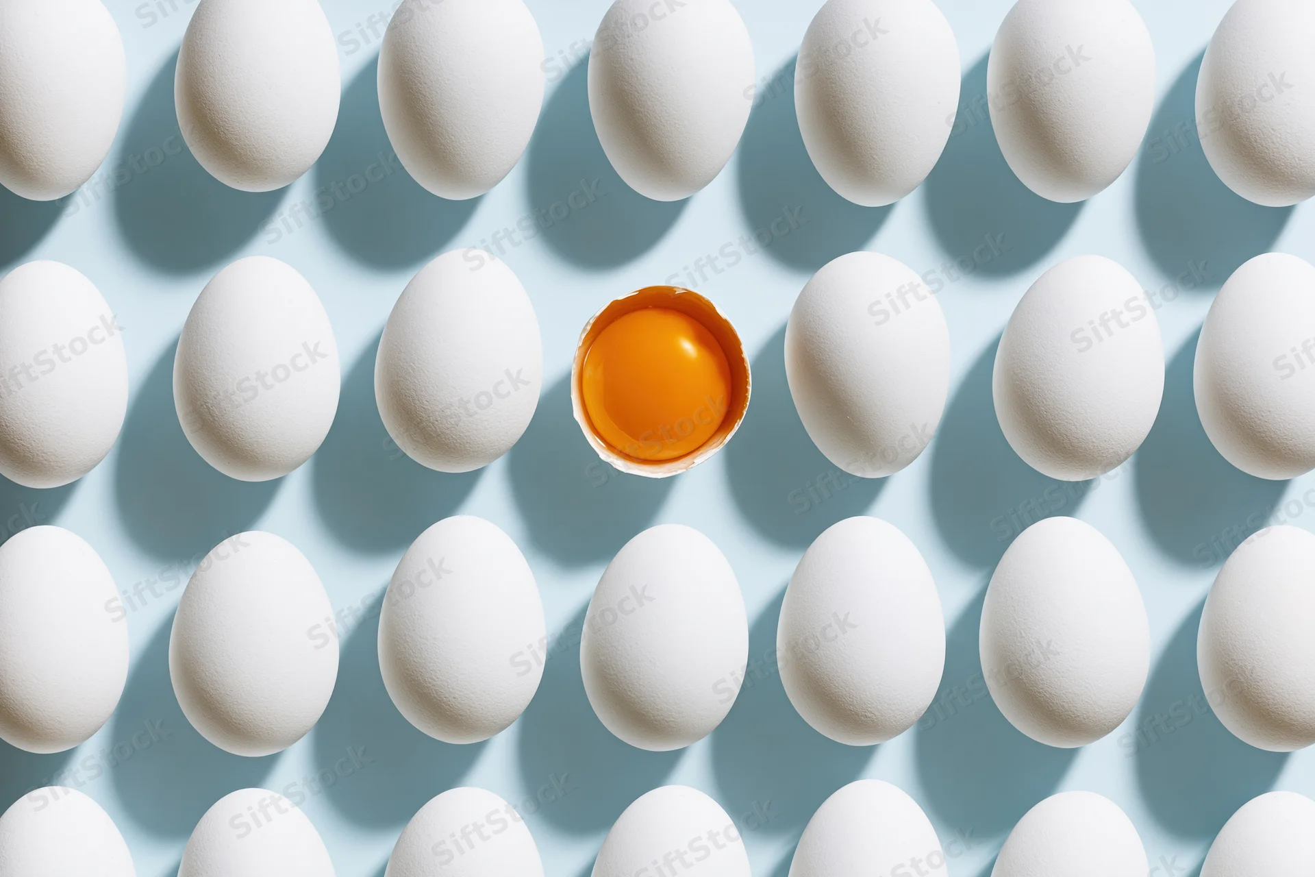 Grid of white eggs arranged in uniform rows on pale blue background with one cracked egg revealing yolk at center