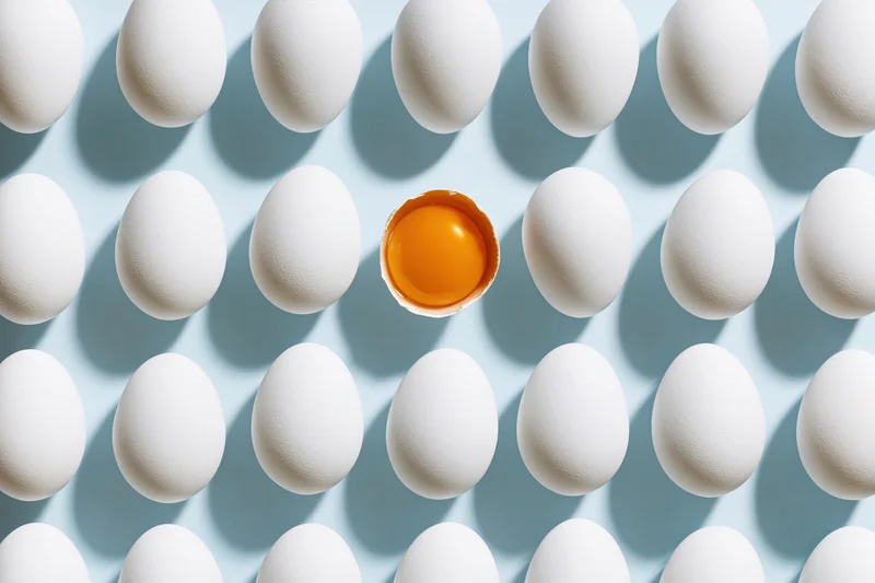 Grid of white eggs arranged in uniform rows on pale blue background with one cracked egg revealing yolk at center