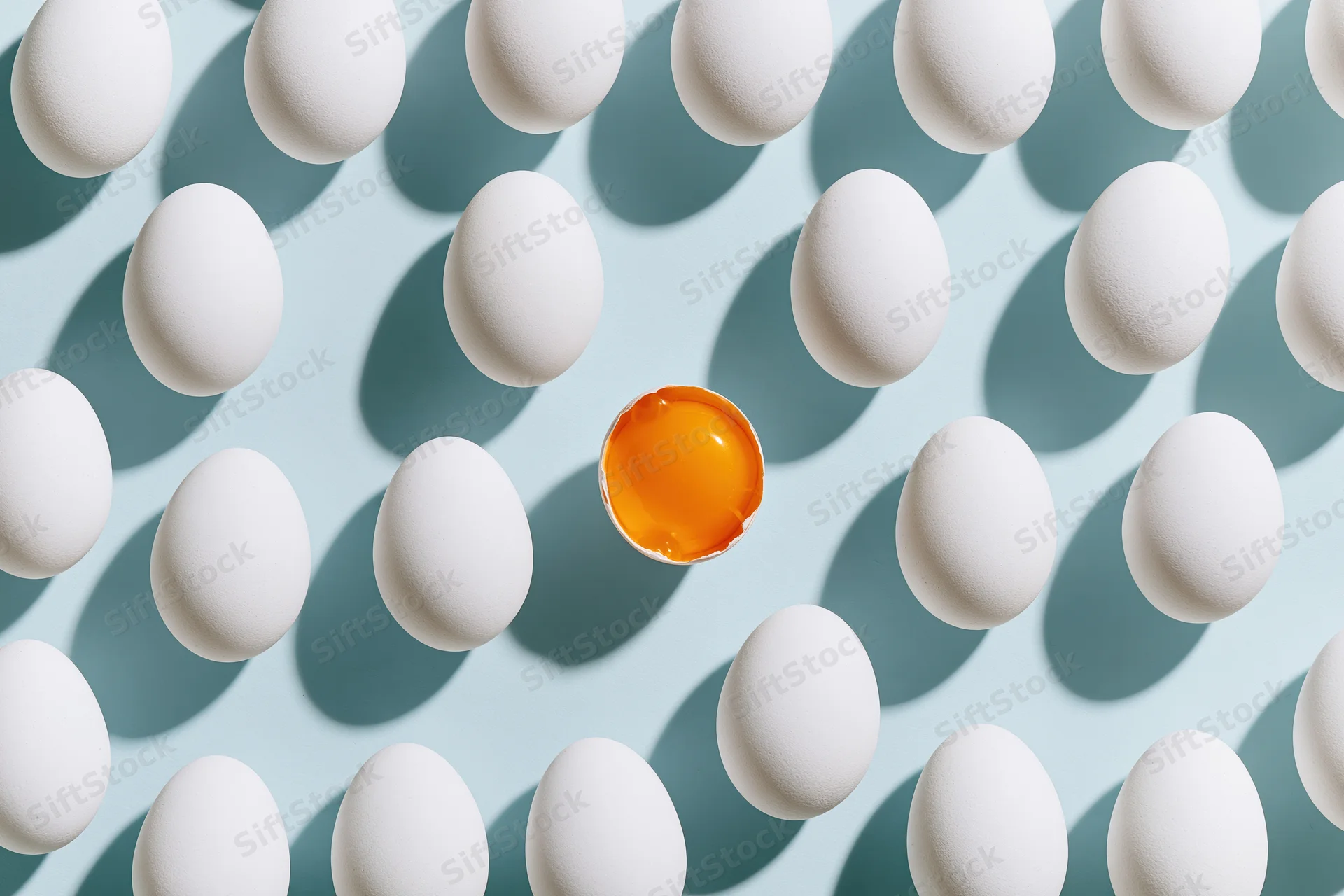Grid of white eggs arranged in uniform rows on pale blue background with one broken egg revealing orange yolk in center