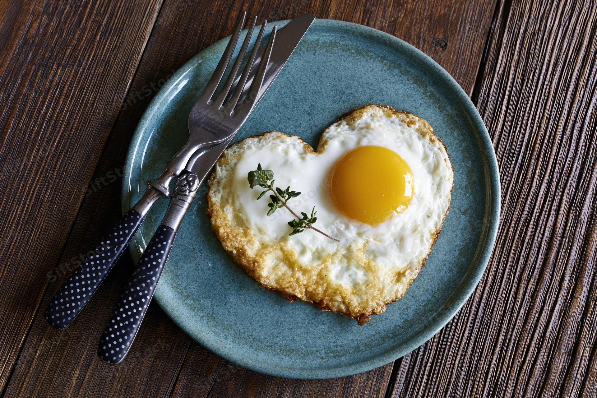 Heart-shaped fried egg with bright yellow yolk on a blue ceramic plate with cutlery on a wooden table