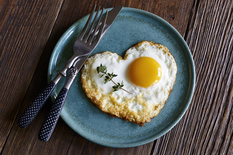 Heart-shaped fried egg with bright yellow yolk on a blue ceramic plate with cutlery on a wooden table