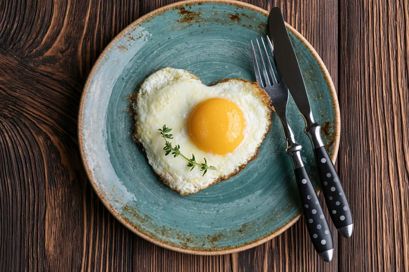 Heart-shaped fried egg with bright yellow yolk on a blue ceramic plate with cutlery on a dark wooden table