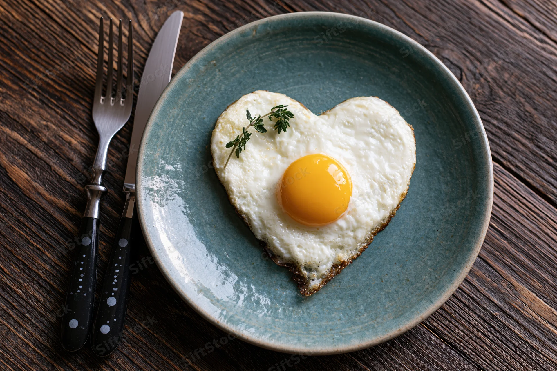 Heart-shaped fried egg with bright yellow yolk on a blue ceramic plate with thyme garnish and cutlery on a wooden table