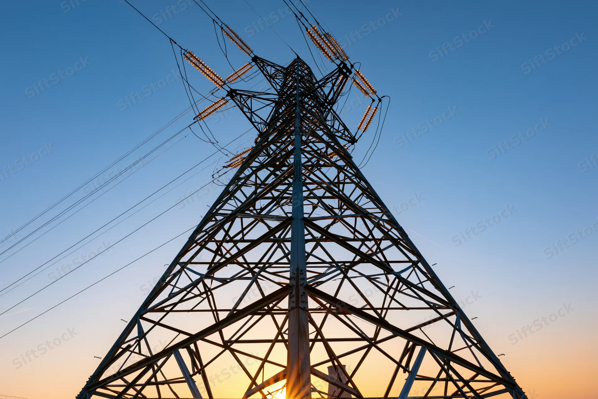High-voltage electricity transmission tower viewed from below against a sunset sky