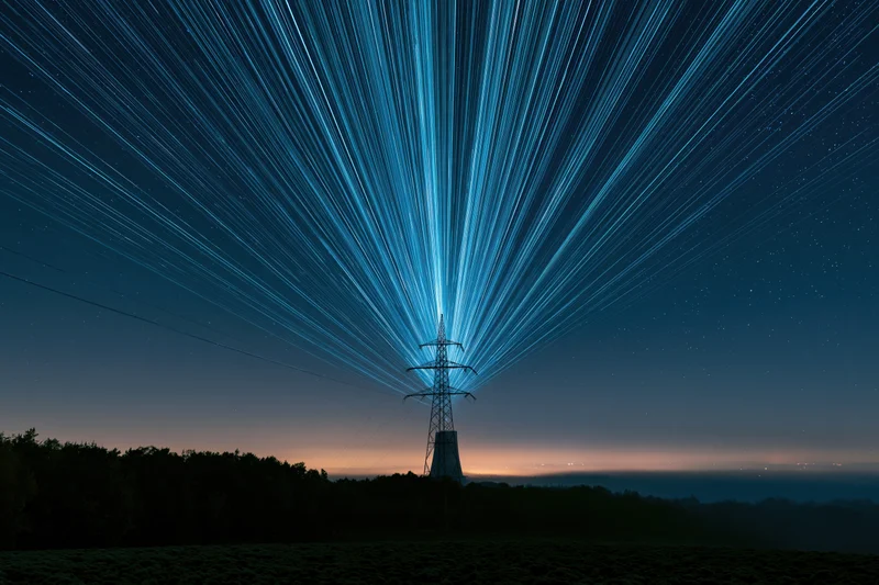High-voltage transmission tower silhouetted against star trails in a rural night landscape