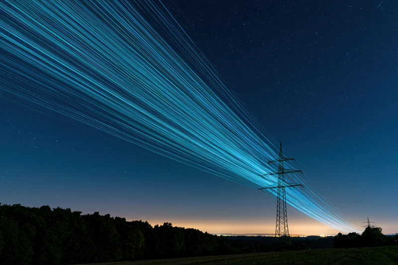 High-voltage transmission tower silhouetted against a starry night sky with blue star trails above a rural landscape