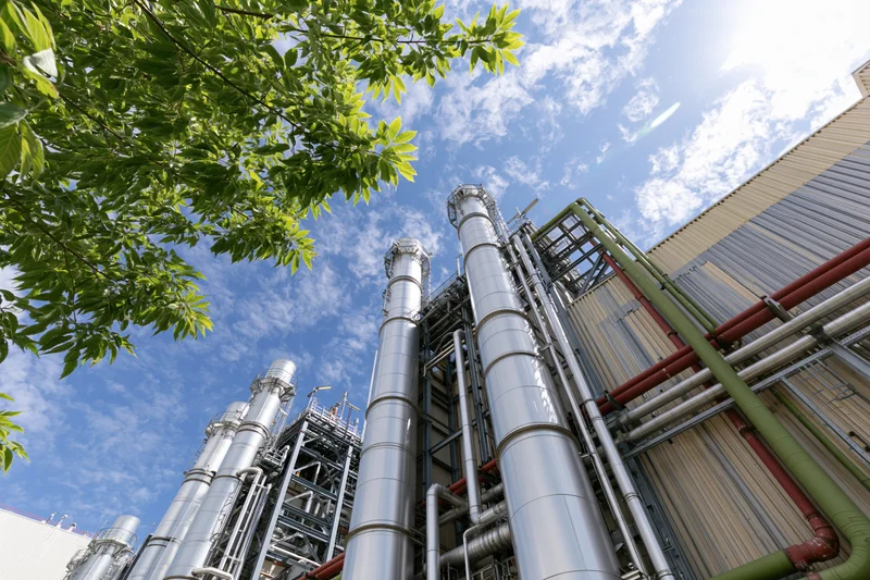Low-angle view of industrial gas turbine power plant with metal pipes and ducts against blue sky and tree foliage