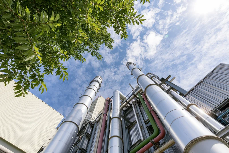 Industrial gas turbine power plant viewed from low angle with pipes and tree foliage against blue sky