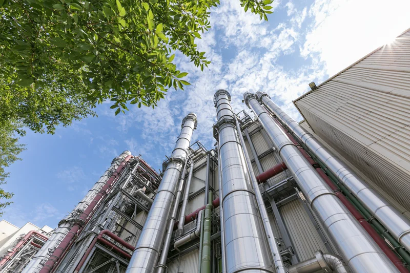 Industrial gas turbine power plant viewed from low angle with metal pipes and chimneys against blue sky and green foliage