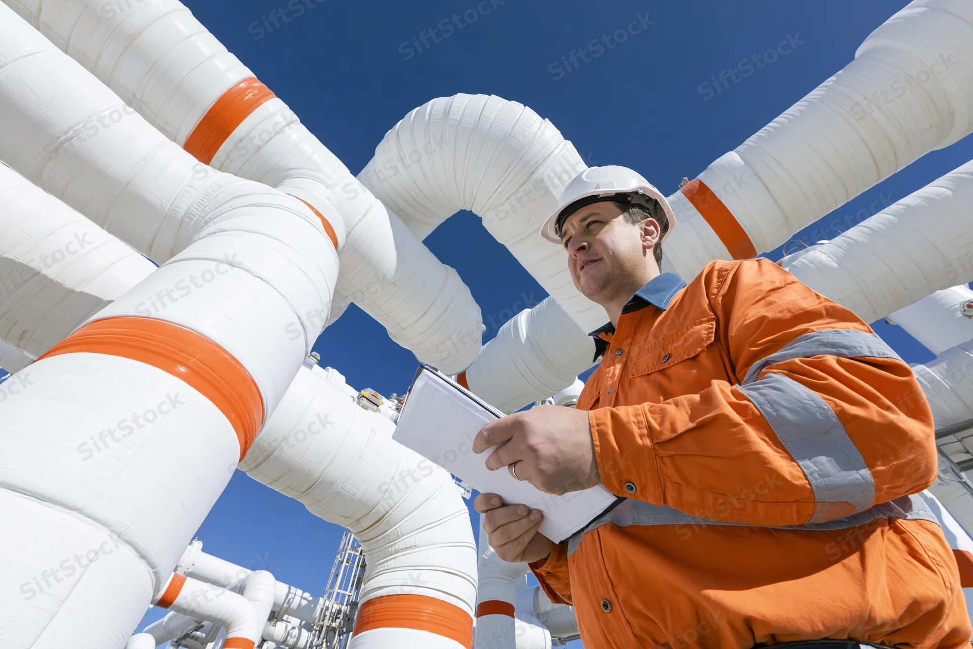 Industrial worker in orange hi-vis jacket and white hard hat inspecting large insulated pipes at a plant site under clear blue sky