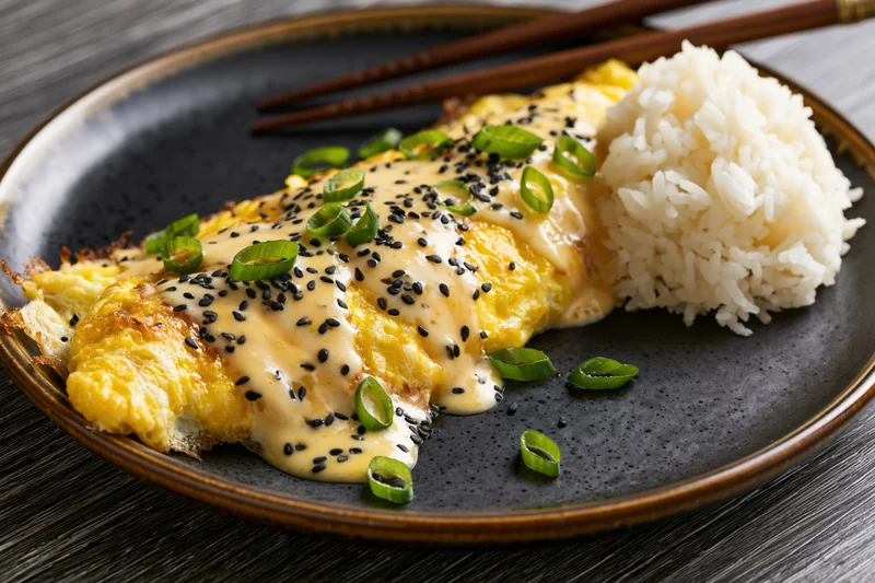 Japanese omurice-style pan-fried egg omelet with golden crisp served on a black ceramic plate with white rice and garnished with green onion and black sesame seeds, chopsticks resting beside it on a wooden table