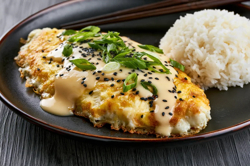 Golden crispy pan-fried egg omelet with white rice and creamy sauce on a black plate