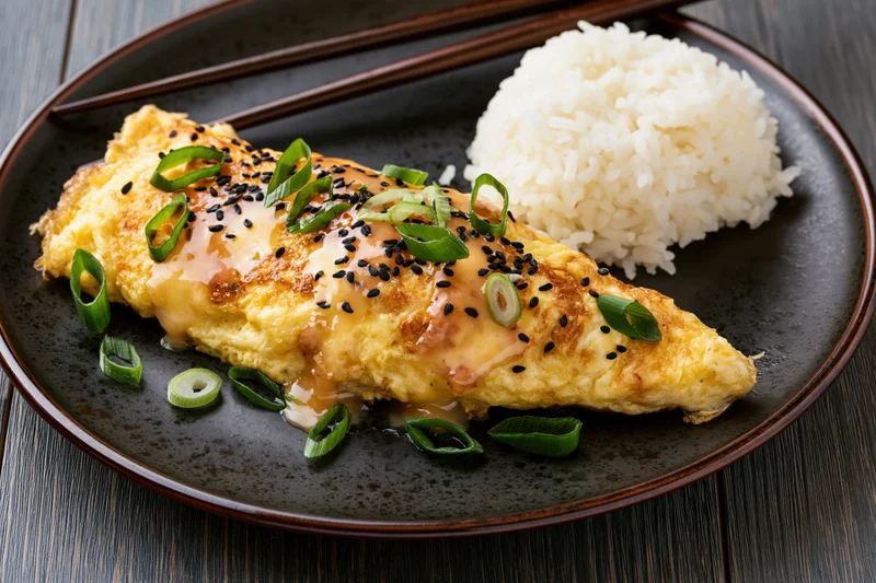 Pan-fried egg omelet with rice and garnishes served on a dark ceramic plate with chopsticks on a wooden table
