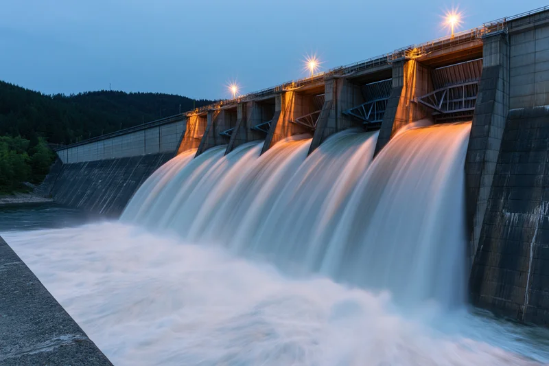 Large concrete hydroelectric dam releasing water through multiple spillways at dusk near forested hills