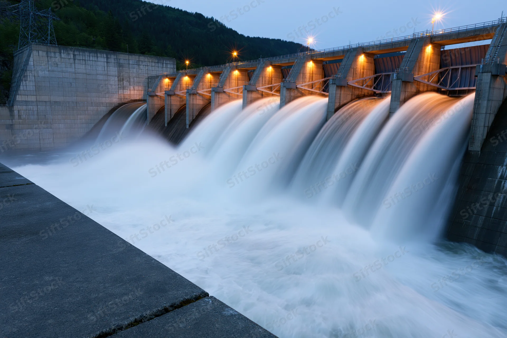 Large concrete hydroelectric dam releasing water through multiple spillways at twilight with illuminated structure and surrounding forested mountains