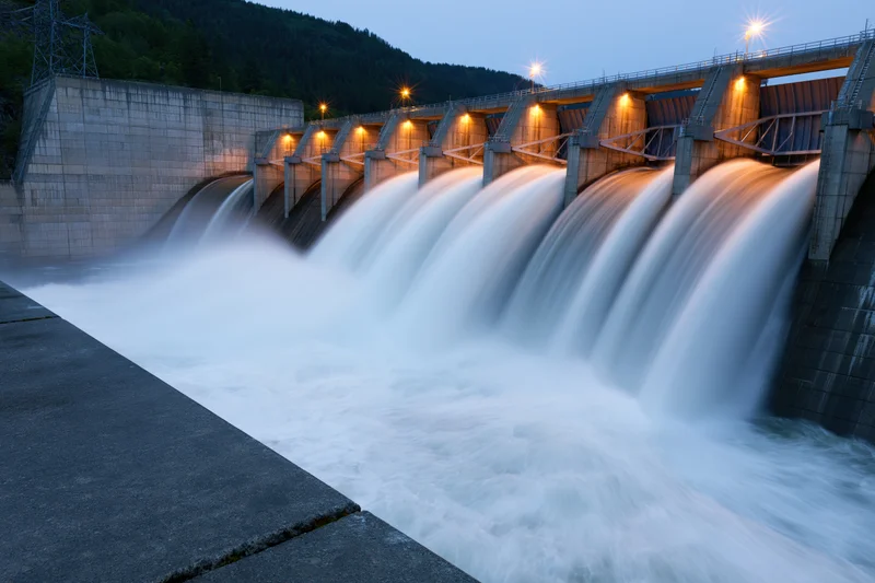 Large concrete hydroelectric dam releasing water through multiple spillways at twilight with illuminated structure and surrounding forested mountains
