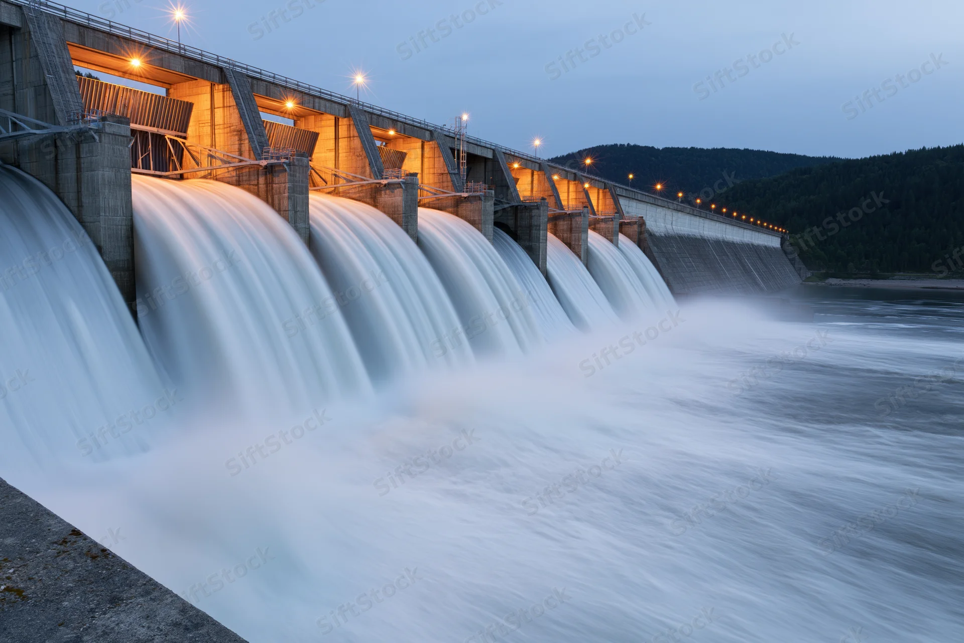 Large concrete hydroelectric dam releasing water through multiple spillways at dusk near forested mountains