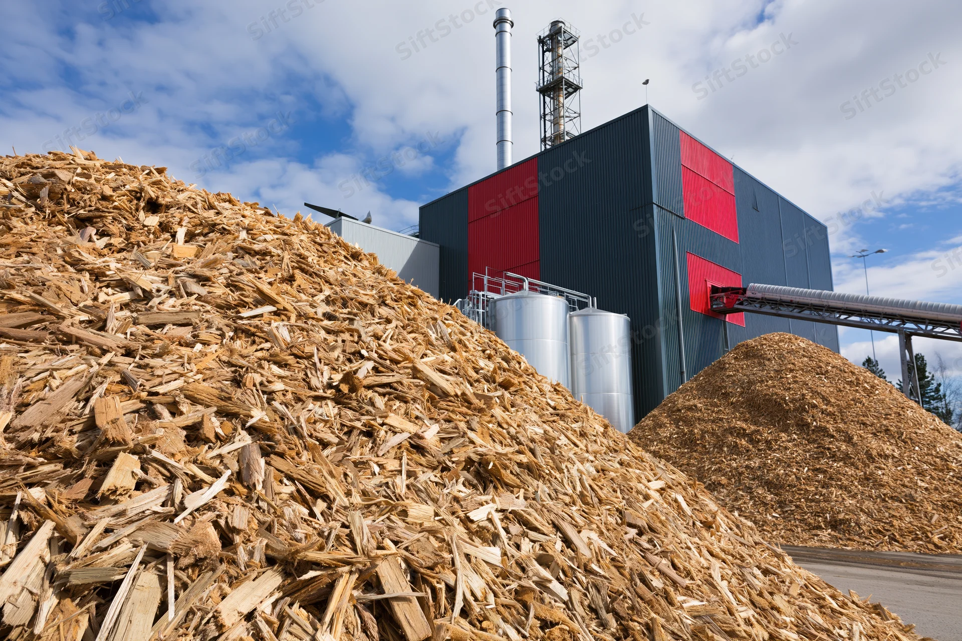 Large mounds of wood chip biomass fuel piled in front of an industrial energy plant with storage silos and a chimney under a partly cloudy sky