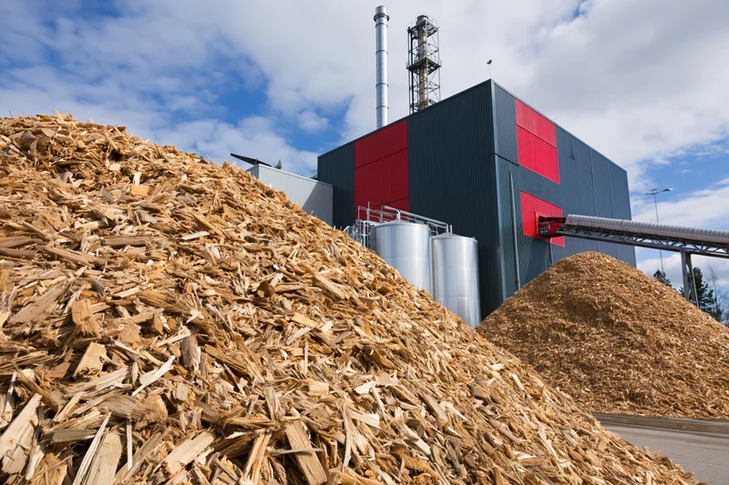 Large mounds of wood chip biomass fuel piled in front of an industrial energy plant with storage silos and a chimney under a partly cloudy sky