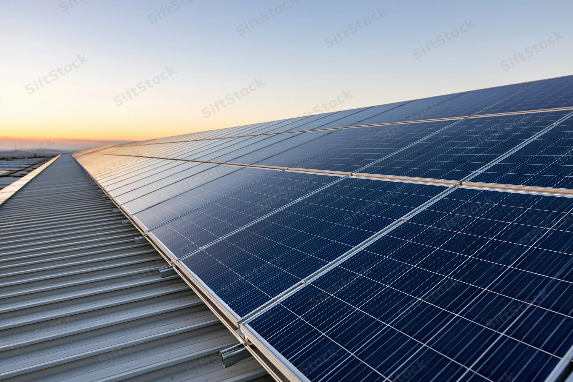 Solar panels arranged in a large array on a metal rooftop stretching toward the horizon at sunset
