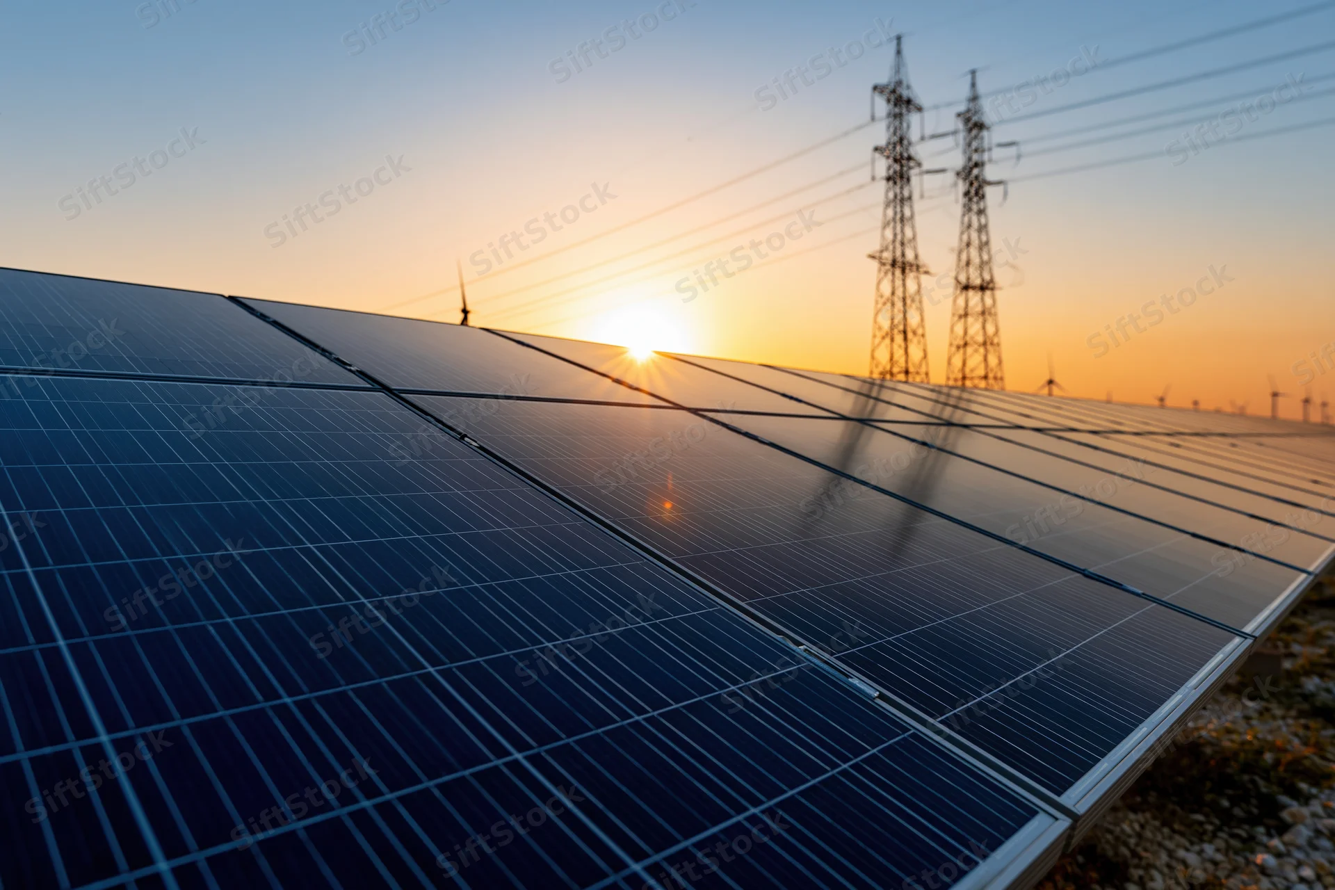 Solar panels arranged in a large array stretching across a field at sunset with power transmission towers in the distance