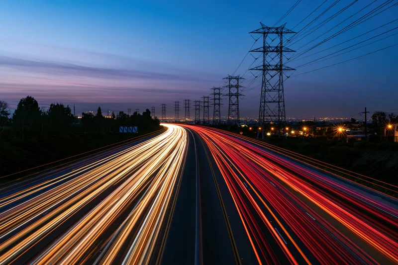 Long-exposure view of a multi-lane highway at dusk with light trails from moving vehicles and power lines along the roadside