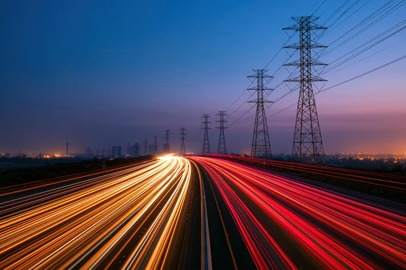 Long-exposure view of a multi-lane highway at dusk with light trails from moving vehicles and power transmission towers alongside