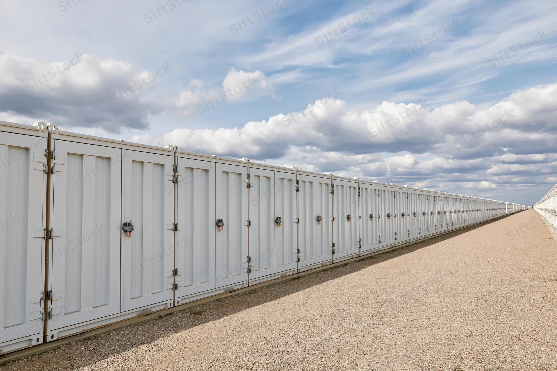 Long row of white industrial battery energy storage units aligned outdoors on gravel ground under blue sky with clouds