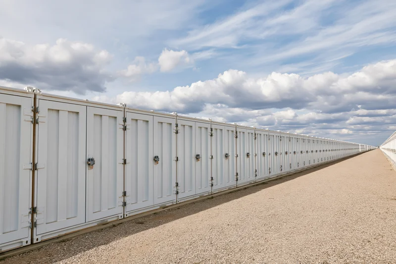 Long row of white industrial battery energy storage units aligned outdoors on gravel ground under blue sky with clouds