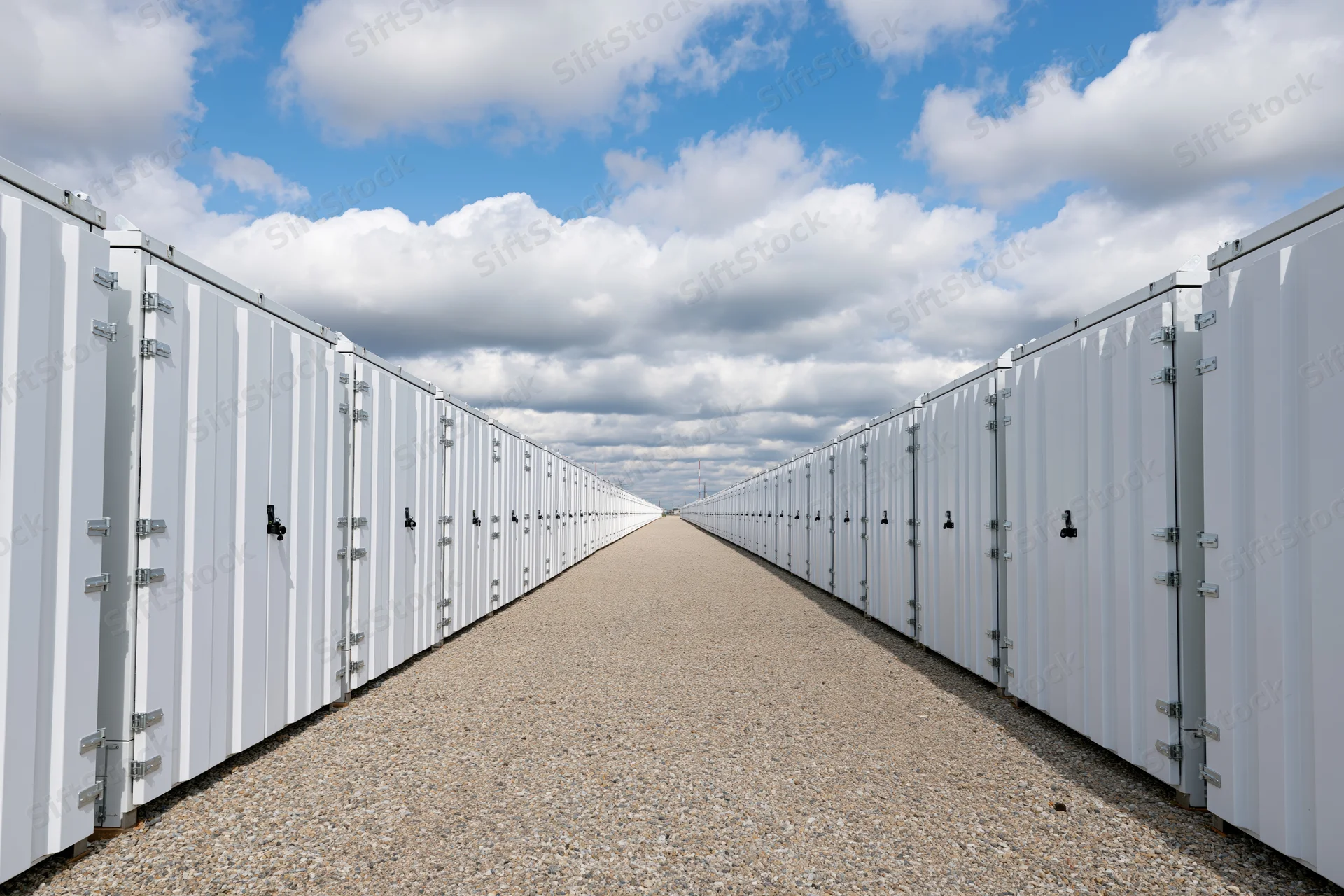Long row of white industrial battery energy storage containers aligned on gravel pathway under blue sky with clouds