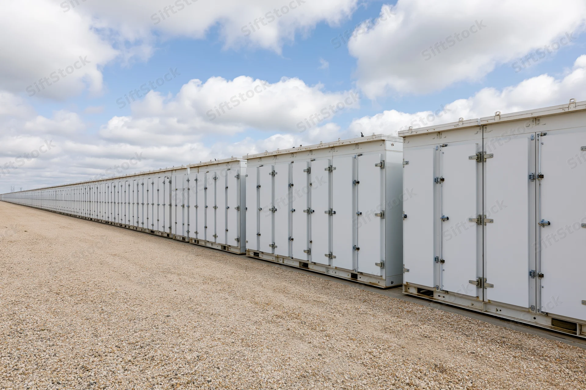 Long row of white industrial battery energy storage units aligned on gravel ground under blue sky with clouds