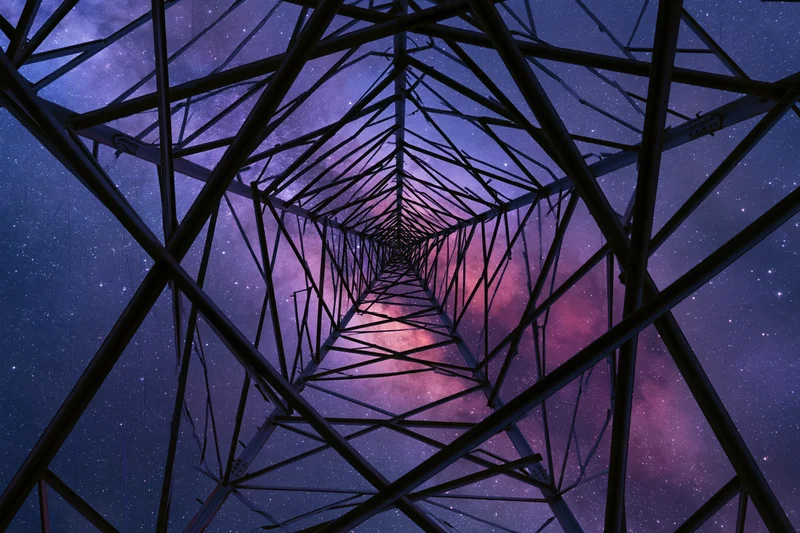 View up through steel lattice of electricity transmission tower at night against starry sky with milky way and nebula