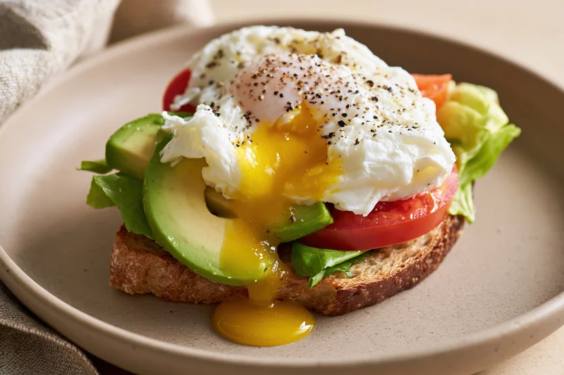 Open-faced breakfast sandwich with poached egg, avocado, tomato and lettuce on toast served on a round ceramic plate