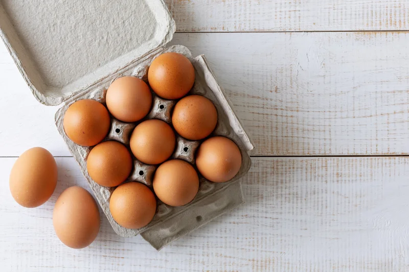 Brown chicken eggs in open cardboard carton on white wooden surface with two eggs beside it