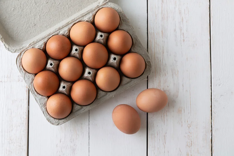 Open cardboard egg carton filled with brown chicken eggs on white wooden surface with two eggs beside it